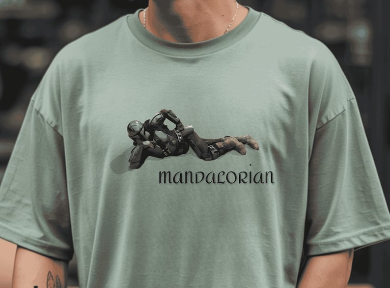 May include: Sage green t-shirt featuring a graphic of a character in black armor, lying down. The word "mandalorian" is printed in a stylized font below the image. The shirt has a crew neck and short sleeves.