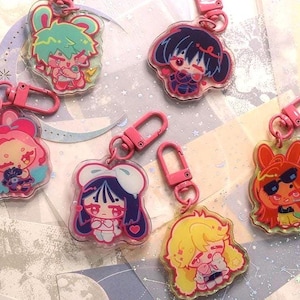 May include: A selection of colourful acrylic keychains with anime-style characters. Each keychain has a unique design with vibrant colours, including pink, blue, yellow, and green. The keychains are attached to pink metal clasps.