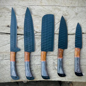 以下が含まれることがあります： 濃い青色の刃と木製の柄が付いた、手作りのキッチンナイフ5本。 刃はテクスチャ加工された表面です。 ハンドルは明るい茶色で、赤いアクセントとグレーと黒のベースが付いています。 ナイフは木製の表面に配置されています。