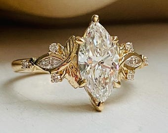 May include: A gold ring featuring a large marquise-cut center stone flanked by smaller marquise and round-cut accent stones. The band includes intricate leaf-like metal detailing extending from the central setting, creating a botanical appearance.