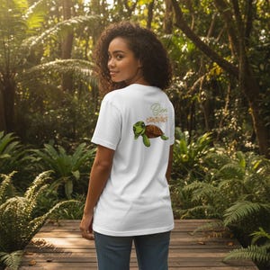 Könnte beinhalten: Weißes T-Shirt mit einem Cartoon-Schildkröten-Motiv und dem Text "Bien Carapace" in Grün und Orange. Die Schildkröte hat einen grünen Kopf und Gliedmaßen sowie einen braunen Panzer. Das Shirt ist aus weichem Material.