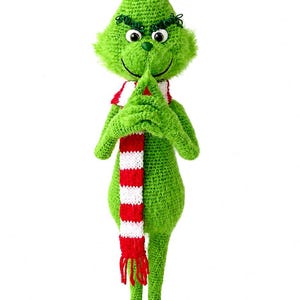 May include: A green crocheted doll with a pointed head, fuzzy texture, and a mischievous facial expression featuring dark eyebrows and a stitched smile. The doll wears a long, red and white striped scarf with fringe at the bottom and has long limbs.