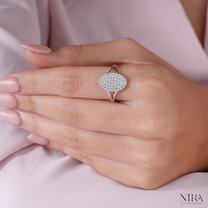 Könnte beinhalten: Ein silberner Ring mit einem ovalen Oberteil, das mit kleinen, funkelnden Steinen besetzt ist. Der Ring befindet sich an einer Hand mit gepflegten Nägeln, vor einem weichen rosa Hintergrund. Das Ringdesign hat ein geteiltes Band.