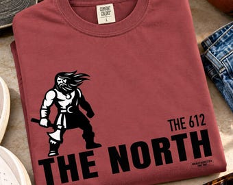 May include: A maroon short-sleeved t-shirt featuring a black and white graphic of a bearded warrior holding an axe. The text "THE 612" and "THE NORTH" is printed in bold black letters below the graphic, with a small "UNAUTHORIZED INK" logo.