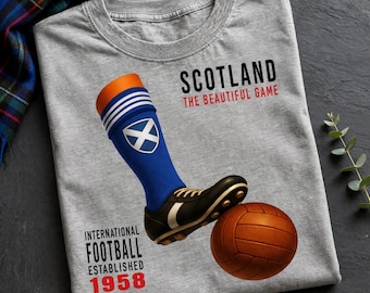 May include: A heather gray t-shirt featuring a graphic of a blue soccer sock with a white saltire emblem, a black cleat, and a brown soccer ball. Text reads SCOTLAND THE BEAUTIFUL GAME and INTERNATIONAL FOOTBALL ESTABLISHED 1958 in black and red.