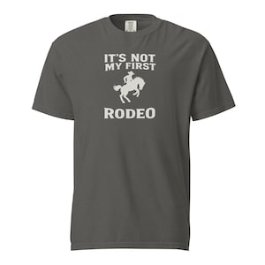 May include: Dark gray t-shirt with the text "IT'S NOT MY FIRST RODEO" in white. The design includes a silhouette of a person riding a horse. The shirt is a crew neck and has short sleeves.