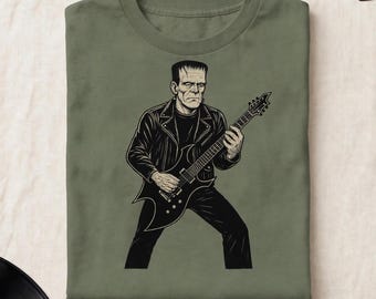 Puede incluir: Una camiseta de color verde oliva con una ilustración gráfica en blanco y negro de una figura que se asemeja al monstruo de Frankenstein, vistiendo una chaqueta de cuero y tocando una guitarra eléctrica, centrada en el pecho de la prenda.