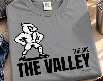 Op de afbeelding: Een grijs t-shirt met ronde hals en een witte afbeelding van een gespierde kardinaal-mascotte in een shirt met het nummer 1. Onder de mascotte staat de tekst THE 602 THE VALLEY in dikke zwarte letters, inclusief een klein Unauthorized Ink logo.