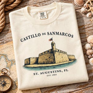 Peut inclure: Un t-shirt à manches courtes de couleur crème avec une illustration du Castillo de San Marcos à St. Augustine, FL. Le motif comprend le texte 'CASTILLO DE SAN MARCOS' au-dessus du fort et 'ST. AUGUSTINE, FL EST. 1565' sous l'illustration.