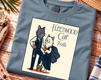May include: A slate blue t-shirt featuring a graphic of two anthropomorphic cats. One cat wears a vest and holds a pocket watch, while the other wears a dark dress. The text reads Fleetwood Cat Trills in a stylized black font on a cream background.