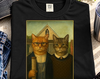 May include: A black t-shirt featuring a graphic print of two cats standing in front of a house. The cat on the left wears glasses and holds a scratching post, while the cat on the right wears a collared shirt. The Comfort Colors brand tag is visible.