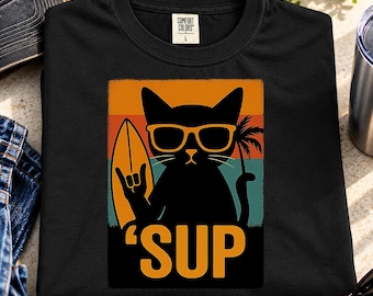 Puede incluir: Una camiseta negra con un gráfico de un gato negro con gafas de sol amarillas, sosteniendo una tabla de surf y haciendo un gesto con la mano. El fondo es un diseño de atardecer con rayas naranjas, amarillas y verde azulado. El texto 'SUP' está impreso en amarillo.