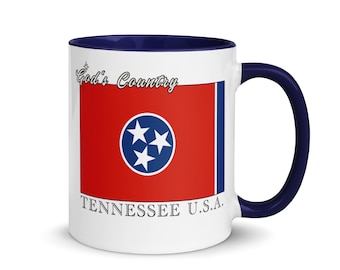 Puede incluir: Taza de cerámica blanca con interior y asa azul marino. La taza presenta el diseño de la bandera del estado de Tennessee, con fondo rojo, un círculo azul con tres estrellas blancas y el texto "God's Country" y "TENNESSEE U.S.A."