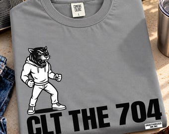 Może przedstawiać: Szary t-shirt z krótkim rękawem z czarno-białą grafiką przedstawiającą panterę w bluzie z kapturem w pozycji walki. Poniżej grafiki znajduje się napis CLT THE 704 pogrubionymi czarnymi literami. Mała biała metka zawiera napis UNAUTHORIZED INK INC.