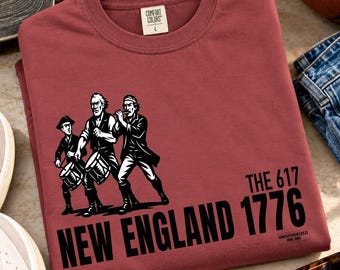 以下が含まれることがあります： レンガ色のクルーネックTシャツ。ドラムとフルートを演奏する3人の人物の黒いグラフィックがプリントされています。テキストは「THE 617 NEW ENGLAND 1776」および「UNAUTHORIZED INK INC」と書かれています。襟の内側にComfort Colorsのラベルがあります。