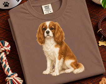 Puede incluir: Una camiseta marrón de cuello redondo con una ilustración gráfica de un Cavalier King Charles Spaniel sentado en el centro. El perro tiene el pelo largo y ondulado de color castaño y blanco. La camiseta tiene una etiqueta de Comfort Colors.