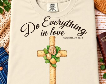 Puede incluir: Una camiseta de color crema con el texto negro 'Do Everything in love' y 'CORINTHIANS 16:14'. Debajo del texto hay un gráfico de una cruz de madera adornada con hojas verdes y tres conchas de color canela. La camiseta tiene una etiqueta Comfort Colors.
