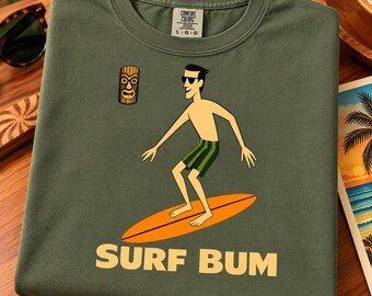 May include: An olive green t-shirt featuring a graphic of a person wearing sunglasses and green striped shorts, standing on an orange surfboard. Above the person is a small tiki mask icon. The text SURF BUM is printed in large yellow letters.