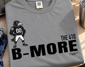 Puede incluir: Una camiseta gris de cuello redondo con un gráfico en blanco y negro de un cuervo con uniforme de fútbol americano y el número 00. El texto dice THE 410 B-MORE y UNAUTHORIZED INK INC. La camiseta tiene una etiqueta de Comfort Colors.