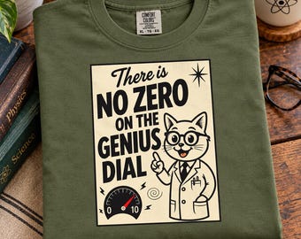 May include: An olive green t-shirt featuring a graphic of a cat wearing glasses and a lab coat. The text reads, There is NO ZERO ON THE GENIUS DIAL, accompanied by an illustration of a dial gauge with a red needle pointing past zero.