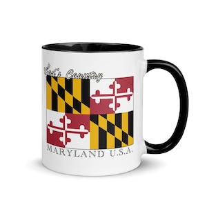 May include: White ceramic mug with a black interior and handle. The mug features the Maryland state flag design, with the words "God's Country" and "Maryland U.S.A." printed on the side. The flag design includes black and gold diagonal stripes and red and white crosses.