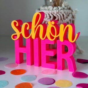 Könnte beinhalten: Ein neonpinkes und gelbes Schild mit der Aufschrift "Schön HIER". Das Wort "Schön" ist in gelber Schrift, und "HIER" ist in Blockbuchstaben. Das Schild steht auf einer weißen Oberfläche mit bunten Konfetti und einer Vase mit getrockneten Pflanzen im Hintergrund.