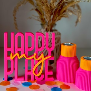 Könnte beinhalten: Ein leuchtend pinkfarbenes 3D-gedrucktes Schild mit dem Text "HAPPY my PLACE" in pinken und gelben Buchstaben, begleitet von zwei gerippten, zylindrischen Kerzenhaltern in leuchtendem Pink und Orange, verstreut mit buntem Papierkonfetti.