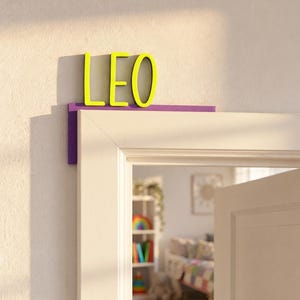 Könnte beinhalten: Ein Tür-Eckschild mit dem Namen LEO in leuchtend gelben Großbuchstaben. Die Buchstaben sind auf einer violetten L-förmigen Basis montiert, die so konzipiert ist, dass sie passgenau auf die obere Ecke eines Türrahmens als Dekoration aufgesetzt wird.