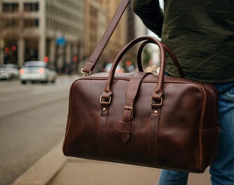 May include: A brown leather duffel bag with a shoulder strap and top handles. The bag has metal buckles and a rectangular shape. The bag is being carried on a city street.