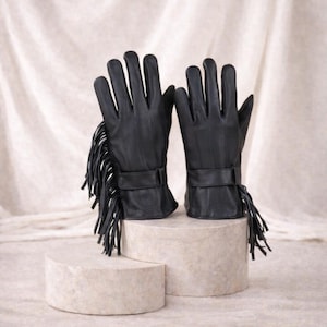 May include: A pair of black leather gloves with fringe detailing. The gloves are displayed on a neutral-toned surface, showcasing their sleek design and texture. Each glove features a strap and fringe accents, adding a touch of style.