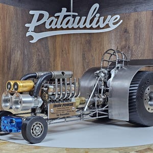 Op de afbeelding: Gedetailleerde metalen sculptuur van een dragster met een grote achterband. De sculptuur heeft een complex motorontwerp met gouden en zilveren componenten, een rolkooi en de tekst "IRON POWER". Het bord boven de dragster luidt "Pataulive".