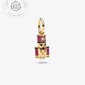 Peut inclure: Un charm doré en forme de casse-noisette soldat. Le charm a un chapeau et des manchettes rouges et roses scintillants. Une petite gemme en forme d'étoile est sur le chapeau. Le charm a des bottes blanches et des accents noirs.