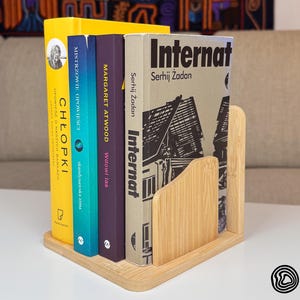May include: A wooden book holder with four books of different colours. The books are titled "Chłopi", "Mistrzowie Opowieści", "Wdowi Las", and "Internat". The book holder is made of light-coloured wood and has a triangular shape.