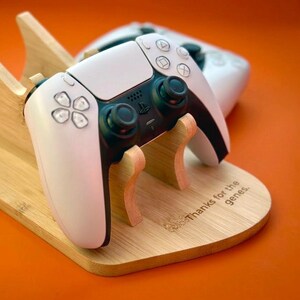 May include: A light-coloured wooden controller stand with two curved supports holding a white and black game controller. The base features the engraved text Thanks for the genes. A small purple heart-shaped object sits to the left of the stand.