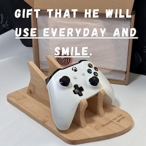 Può includere: Un supporto per controller in legno chiaro che sostiene un controller di gioco Xbox bianco. Il supporto presenta il testo inciso 'Thanks for the genes.' Sullo sfondo, una scatola di cartone aperta mostra il testo 'GIFT THAT HE WILL USE EVERYDAY AND SMILE.'