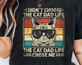May include: Black t-shirt featuring a cat wearing sunglasses, with the text "I Didn't Choose The Cat Dad Life, The Cat Dad Life Chose Me." The design includes a retro color block background of red, orange, yellow, and teal.
