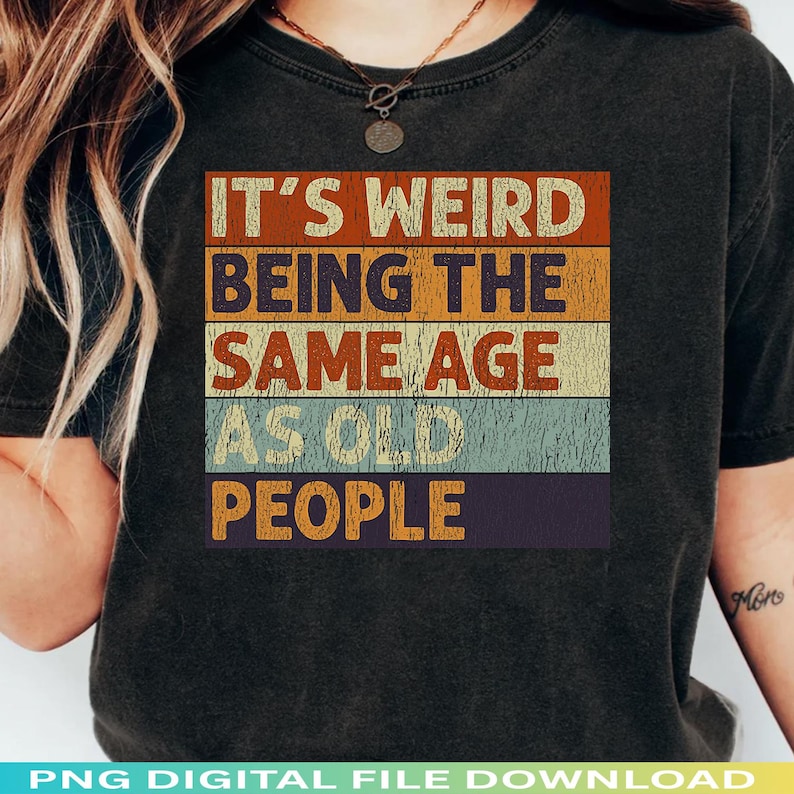 Może przedstawiać: Czarny t-shirt z napisem "IT'S WEIRD BEING THE SAME AGE AS OLD PEOPLE" w stylu retro. Tekst umieszczono w poziomych pasach w kolorach pomarańczowym, ż&oacute;łtym, niebieskim i fioletowym.