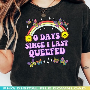 May include: Dark gray t-shirt with a rainbow, smiling suns, and butterflies. The text reads "0 DAYS SINCE I LAST QUEEFED" in a retro font. The shirt has a vintage, worn look.
