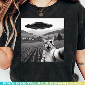 May include: Black t-shirt featuring a black and white graphic of a cat taking a selfie with a UFO hovering above a rural landscape. The cat has wide eyes and an open mouth. The text "PNG DIGITAL FILE DOWNLOAD" is at the bottom.
