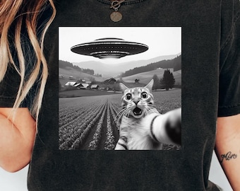May include: Black t-shirt featuring a black and white graphic of a cat taking a selfie with a UFO hovering above a rural landscape. The cat has wide eyes and an open mouth. The text "PNG DIGITAL FILE DOWNLOAD" is at the bottom.