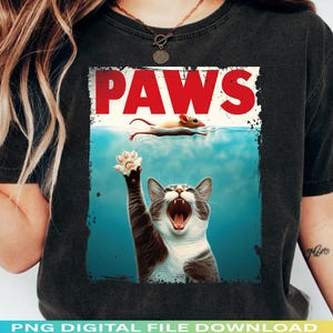 Op de afbeelding: Zwart T-shirt met een afbeelding van een kat die naar een muis grijpt die in het water zwemt. Het woord "PAWS" staat in rode blokletters boven de scène. De kat is grijs en wit met zijn mond open.