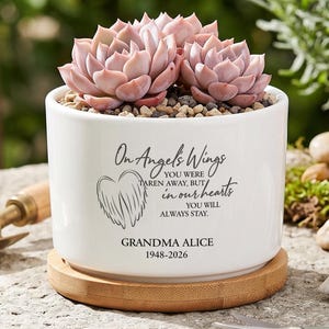 Op de afbeelding: Een witte keramische plantenpot met bamboe schotel en roze vetplanten. De pot is voorzien van een zwarte afbeelding van engelvleugels en de tekst: On Angel's Wings YOU WERE TAKEN AWAY, BUT in our hearts YOU WILL ALWAYS STAY. GRANDMA ALICE 1948-2026.