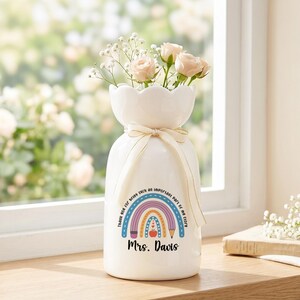 以下が含まれることがあります： 縁がスカラップ状になった白い陶器製の花瓶で、首元にはクリーム色のリボンが結ばれています。花瓶には鉛筆とリンゴが描かれたカラフルな虹のグラフィックと、「Thank you for being such an important part of my story」および「Mrs. Davis」という文字がプリントされています。