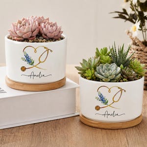 May include: Two white cylindrical ceramic planters with bamboo saucers, each containing assorted succulents. The planters feature a gold stethoscope graphic shaped like a heart with blue flowers and the name Amelia printed in black script.