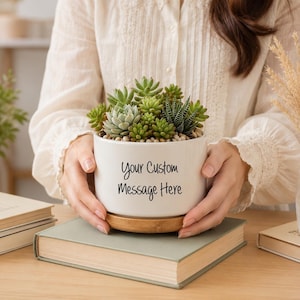 Puede incluir: Una maceta cilíndrica de cerámica blanca con base de bambú, llena de una variedad de suculentas verdes. El frente de la maceta presenta el texto impreso en negro "Your Custom Message Here" en una tipografía sencilla y sin remates.