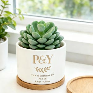May include: A white ceramic cylindrical planter containing a green succulent plant. The planter features gold text reading P&Y, a leaf graphic, and the words THE WEDDING OF PETER AND YARA. It sits on a circular wooden base with a matching wooden lid nearby.