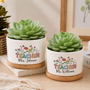 Può includere: Due vasi cilindrici in ceramica bianca con sottovasi in bambù, contenenti ciascuno una pianta succulenta verde. I vasi presentano una grafica floreale colorata con farfalle e il testo "TEACHER" seguito da "Mrs. Johnson" o "Ms. Williams" in corsivo.