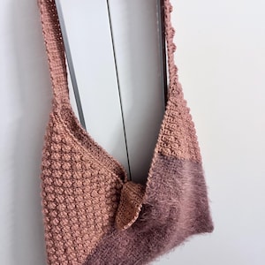 May include: A handmade, dusky pink crochet bag. One side features a textured bobble stitch, the other a fuzzy mauve panel. It has a long strap and a small, knotted closure. The bag is a unique, handcrafted item.