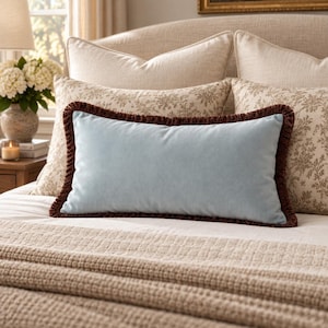May include: A rectangular light blue velvet throw pillow featuring a dark brown fringe trim along all four edges. The pillow is positioned horizontally in the centre of a bed, resting against larger beige floral patterned and solid pillows.