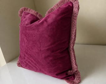 Könnte beinhalten: Ein quadratisches Zierkissen aus tief magenta-farbenem Samtstoff. Die Kanten des Kissens sind mit einer passenden magenta-farbenen Fransenborte versehen, die entlang des gesamten Umfangs verläuft und einen strukturierten Rand bildet.